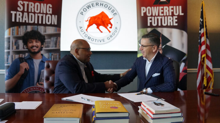 Two individuals shake hands at a table during a formal meeting, with banners displaying “Strong Tradition” and “Powerful Future” and the logo of Powerhouse Automotive Group behind them.